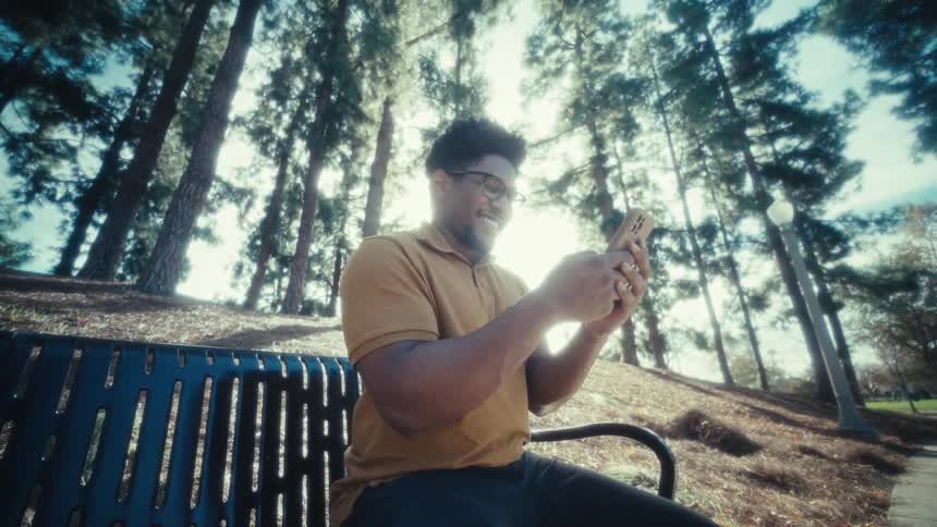 Man seated on a park bench uses a smartphone among trees with sunlight in the background