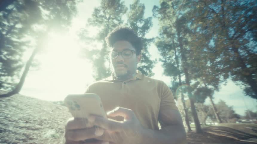 A smiling man walks through a sunny park with tall trees while pulling a smartphone out of his pocket to type a message