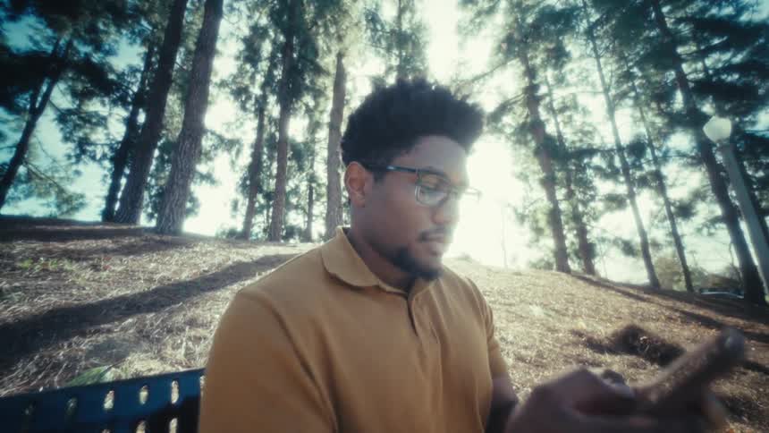 A man with glasses and a beard sits on a park bench surrounded by tall trees and smiles while using his phone