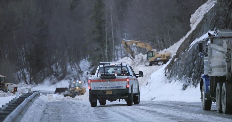 A maintenance pickup truck with flashing lights is stopped on a snowy mountain road as construction equipment works to clear a distant avalanche