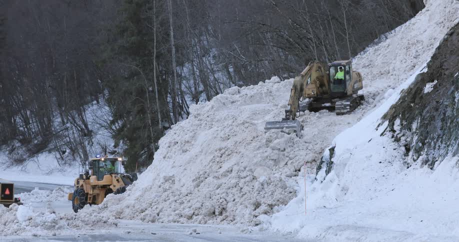 Excavator and wheel loader remove a large buildup of snow along a road in a mountainous area