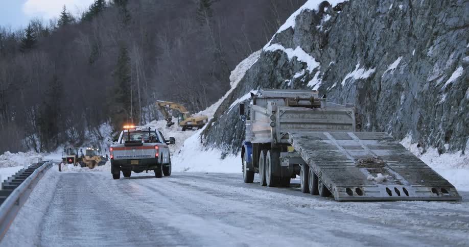Flatbed truck stopped on a snowy road with a police vehicle and construction equipment in the background