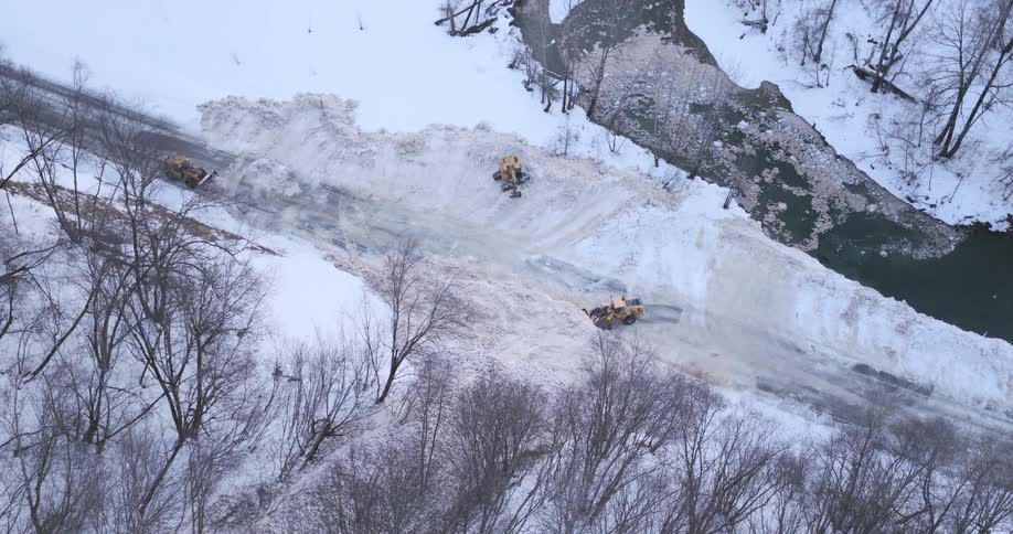An aerial view shows three yellow construction vehicles clearing a massive pile of snow from a road next to a river and bare trees