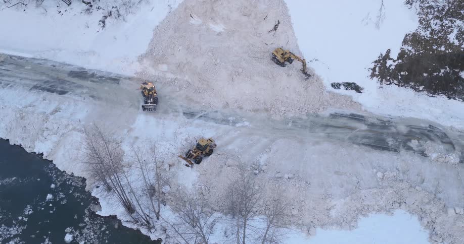 An aerial view shows three yellow construction vehicles clearing a massive pile of snow from a road next to a river and bare trees