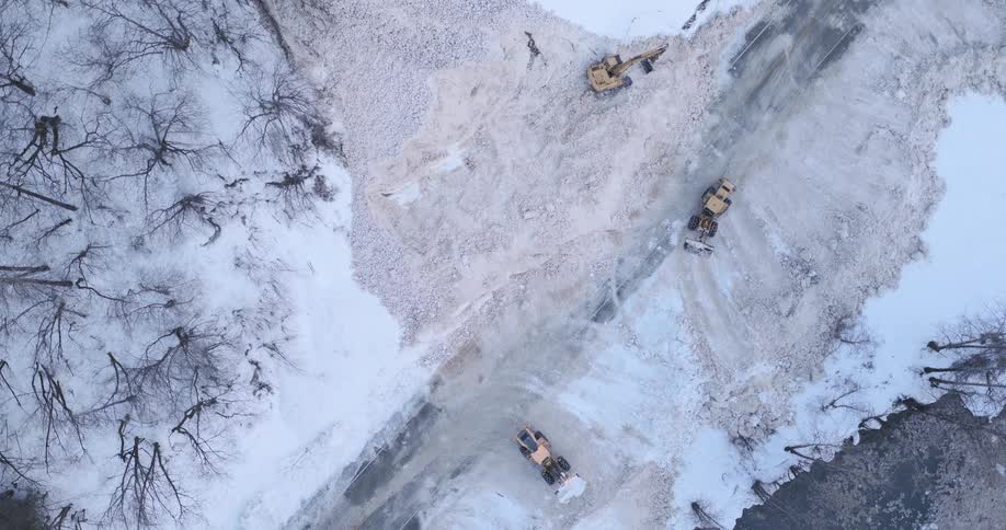 This aerial footage captures multiple pieces of heavy machinery working to clear a massive blockage of white debris from a road located alongside a river a front loader and an excavator are seen actively pushing and maneuvering the material to restore ac