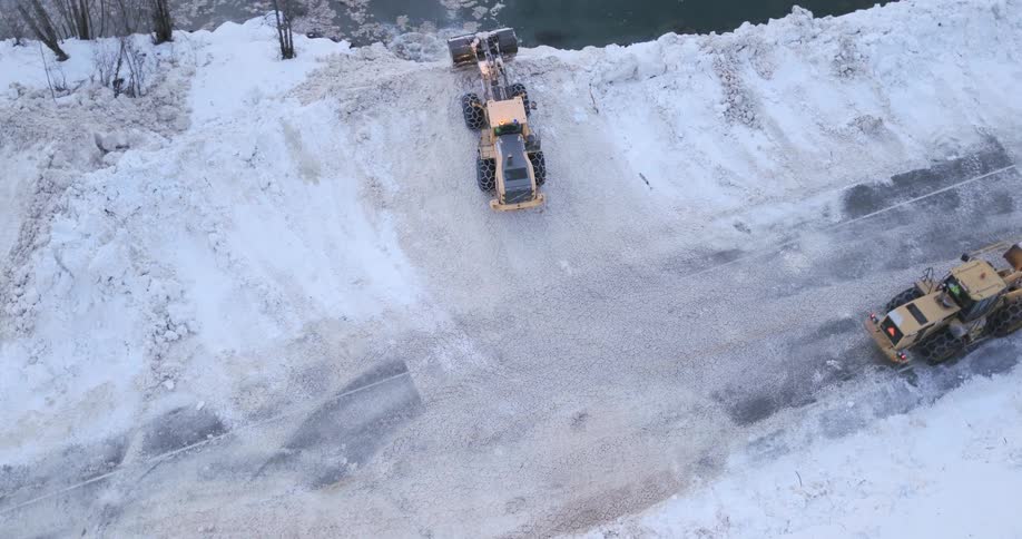 An aerial view shows a large yellow front end loader clearing deep snow and debris from a road next to a frozen river