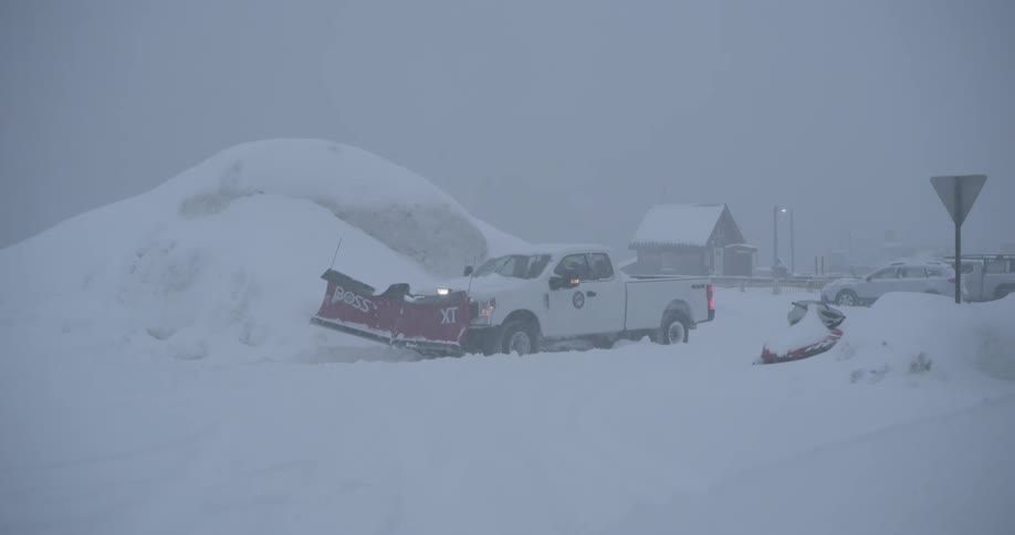 A white pickup truck with a red snowplow clears heavy snow from a country road during a winter storm