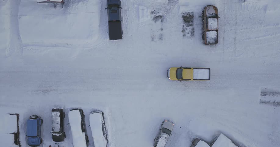 From a topdown aerial perspective a bright yellow pickup truck drives across a parking lot completely covered in a thick blanket of snow passing other stationary vehicles