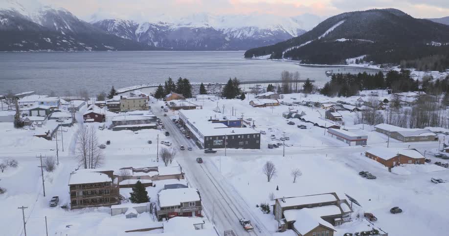 An aerial view shows a small coastal town blanketed in deep snow with a car driving down a central road toward a large body of waterjagged snowcovered mountains rise sharply in the background under a pale wintry sky framing the quiet residential