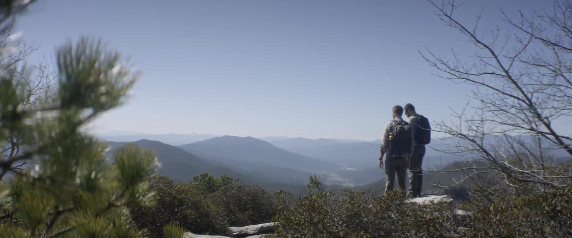 Two hikers stand on a rock overlooking the hills and valley below