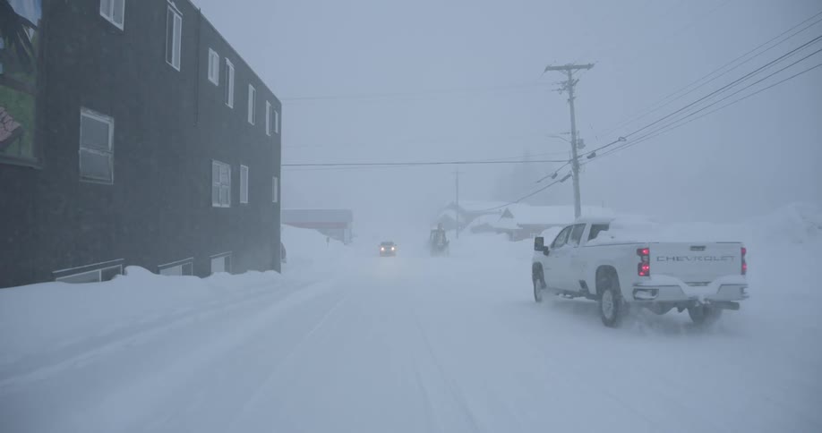 A white pickup truck drives through a heavy snowstorm on a street lined with tall snowbanks and dark buildings