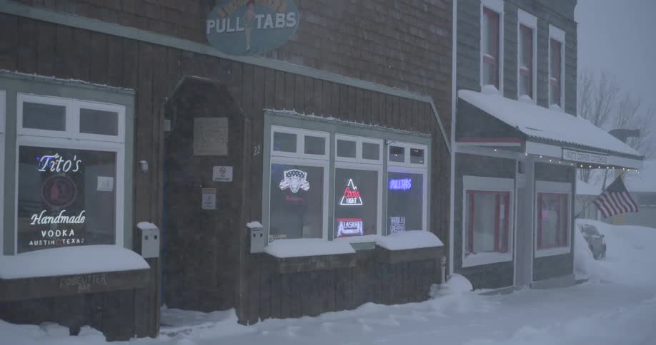 A camera pans across a snowcovered street revealing a rustic wooden building with neon beer signs in the windows dense snowflakes fall steadily against the backdrop of the quiet storefront and an american flag