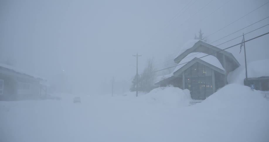 A snowstorm engulfs a quiet rural street with houses barely visible through heavy blowing snow