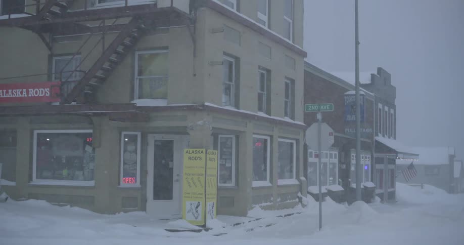 A white pickup truck drives through a heavy snowstorm past local businesses at the corner of main street in a quiet winter town