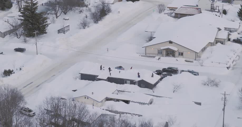 An aerial view shows a snow covered town where a group of people shovel a large flat roof while cars navigate the icy roads below