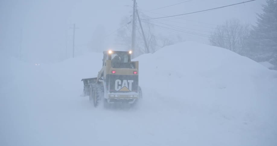 A yellow caterpillar skid steer loader maneuvers through deep snow during a heavy winter blizzardthe vehicles lights are visible through the thick whiteout as it clears a path along a buried residential street