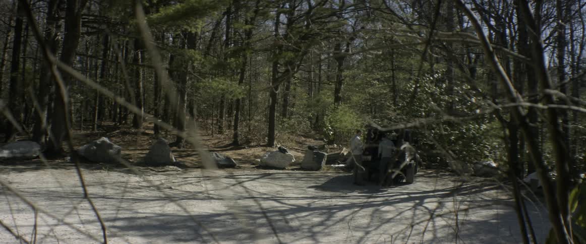 Two men stand at the back of a jeep in a parking lot in a forest