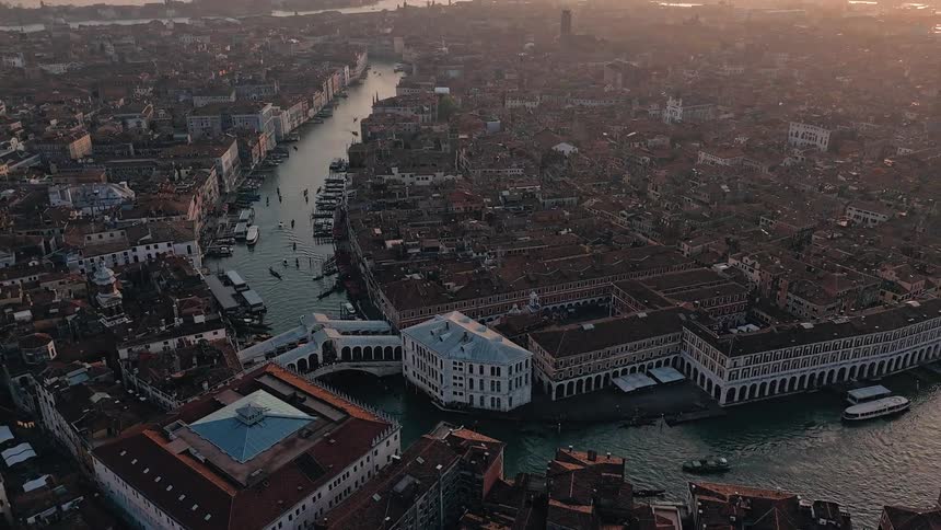 An aerial shot captures the densely packed terracotta rooftops and the bustling grand canal of venice with the rialto bridge at its center during a golden sunset