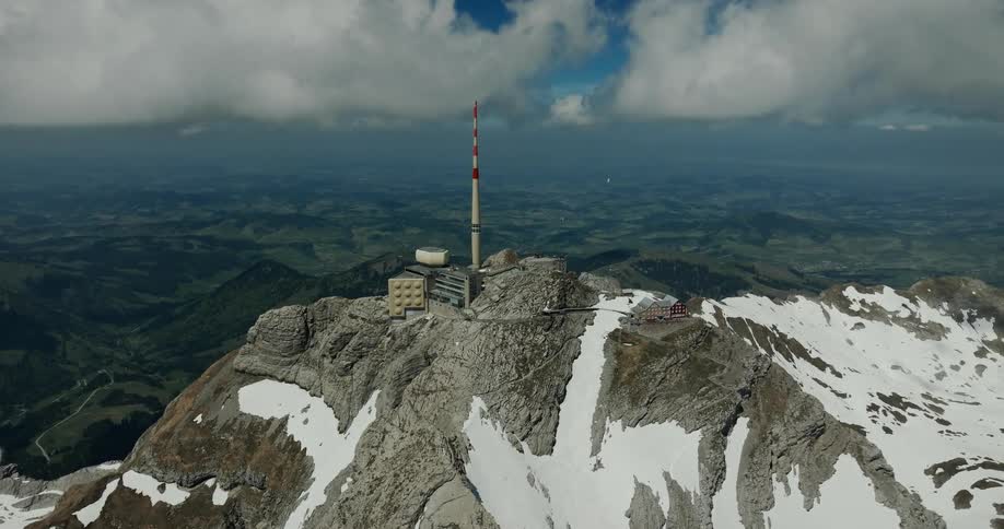 An aerial view shows the snowy peak of a mountain where a tall tower and a few small buildings stand under a cloudy sky