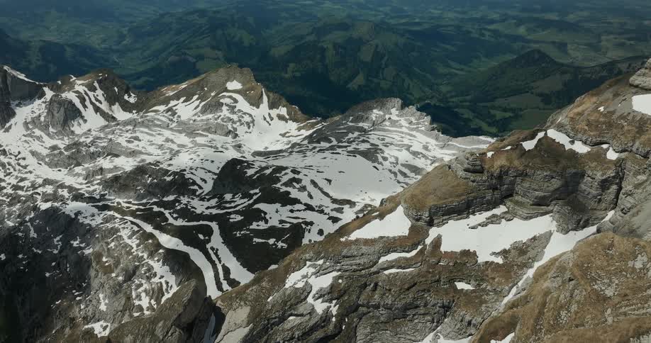 An aerial view shows a vast mountain range with rocky peaks covered in patches of snow above a green valley