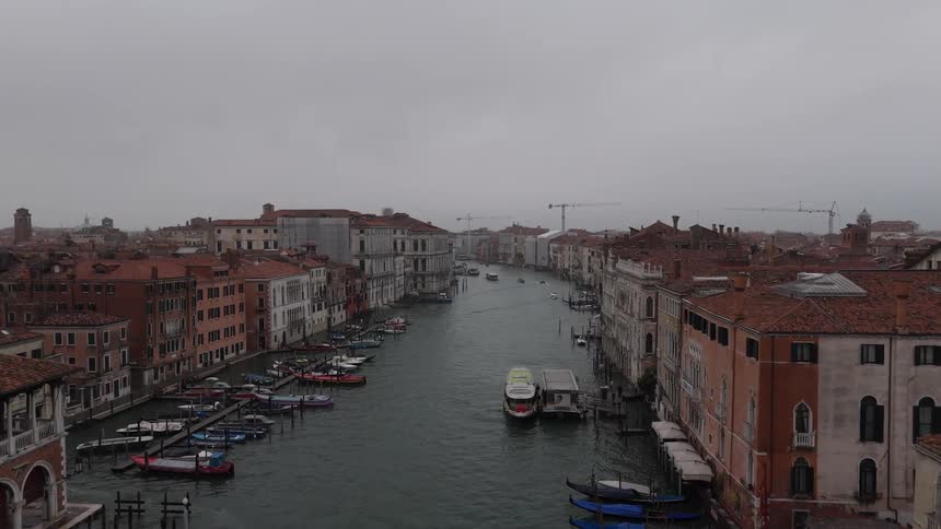 An aerial view shows the grand canal in venice where many boats and gondolas travel between historic buildings under a grey sky