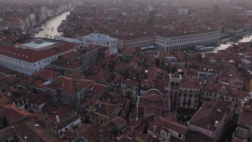 An aerial view pulls back slowly to reveal a historic sprawling cityscape its dense redtiled roofs and winding canals stretching toward a hazy distant horizon at dusk