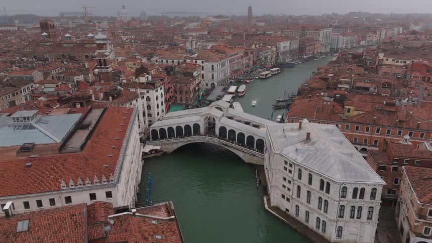 An aerial view shows the rialto bridge crossing the grand canal in venice among many historic buildings and boats under a grey sky