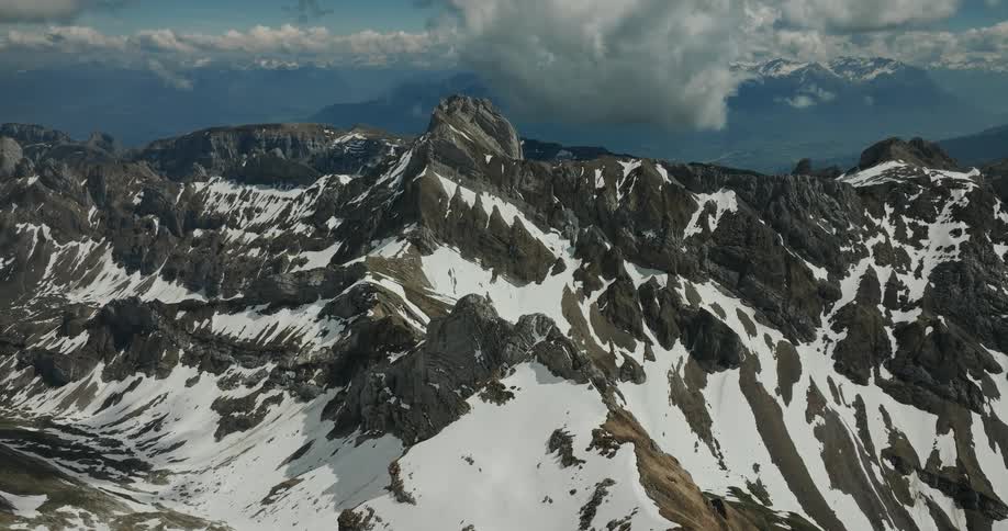 An epic aerial view glides toward a colossal mountain range its jagged rocky peaks rising through blankets of snow under a vast cloudfilled sky