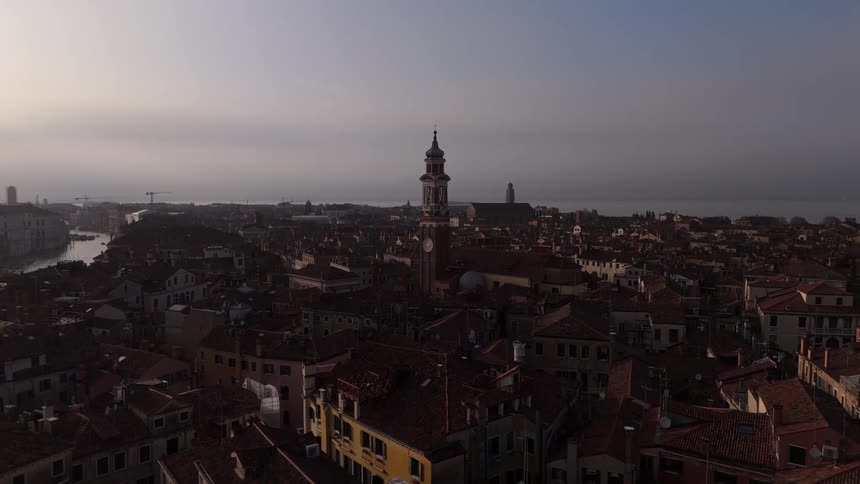 An aerial drone shot glides over the historic rooftops of venice at dawn centered on a prominent bell tower the dim morning light casts long shadows across the dense architecture while the mist settles over the distant lagoon