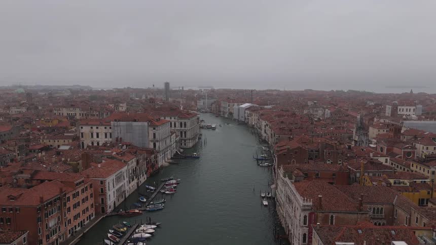 A wide aerial drone shot moves over a european cityscape during a grey overcast day capturing the dense rows of historic terracotta roofed buildings and various small boats docked along the edges of the green water
