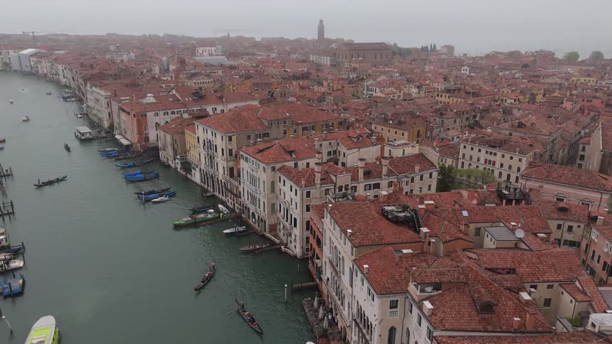A high angle view shows the grand canal in venice with several gondolas and boats moving through the water surrounded by historic buildings with red tile roofs under a grey overcast sky