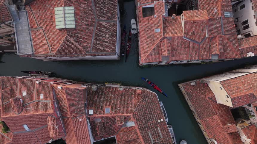 An aerial topdown view shows a narrow canal winding through a dense cluster of buildings with terracotta tile roofsa blue and red gondola glides slowly through the dark water as the camera rotates slightly above the urban waterway