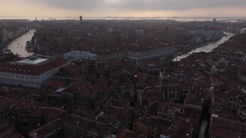 An aerial view shows the dense city of venice with many terracotta rooftops and a winding canal under a hazy sky at sunset