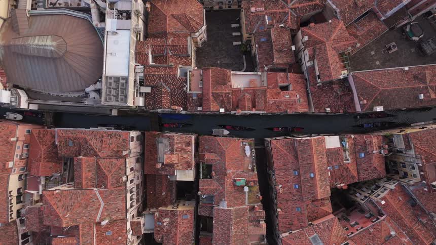 From a stunning topdown aerial view gondolas glide through a narrow canal that cuts a dark line through the dense iconic red rooftops of venice