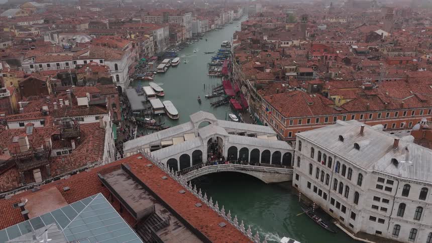 The rooftops of an european city toward a bridge as gondolas and boats navigate under a cloudy sky while tourists walk across the historic stone structure and gather along the nearby docks and walkways