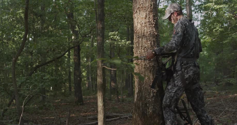 A hunter in camouflage attaches a strap to a tree trunk
