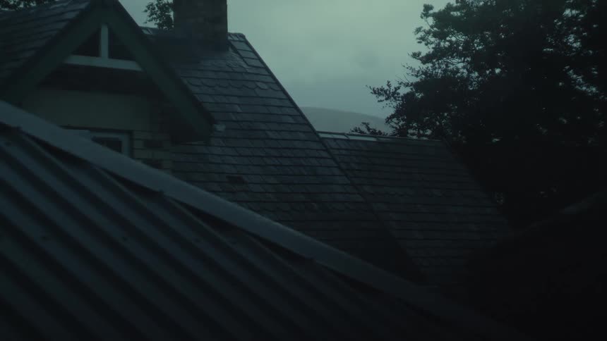 Rain falls on the dark slate roof of a house set against a gloomy overcast sky