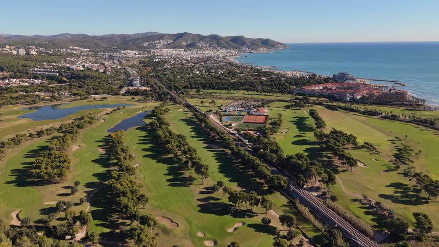 This aerial footage captures a sprawling coastal golf course nestled between the blue mediterranean sea and a dense urban landscapea long straight road and a parallel railway line bisect the lush green fairways leading the eye toward a distant seasid