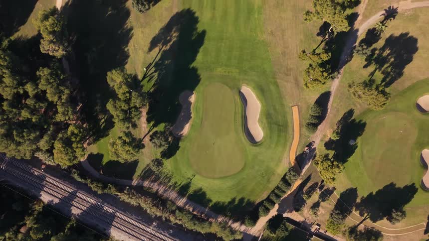 A top down drone shot glides over a green golf course featuring sand traps and fairways crossing over a set of parallel railroad tracks while two golfers walk across the grass during a sunny day with long shadows cast by the surrounding trees