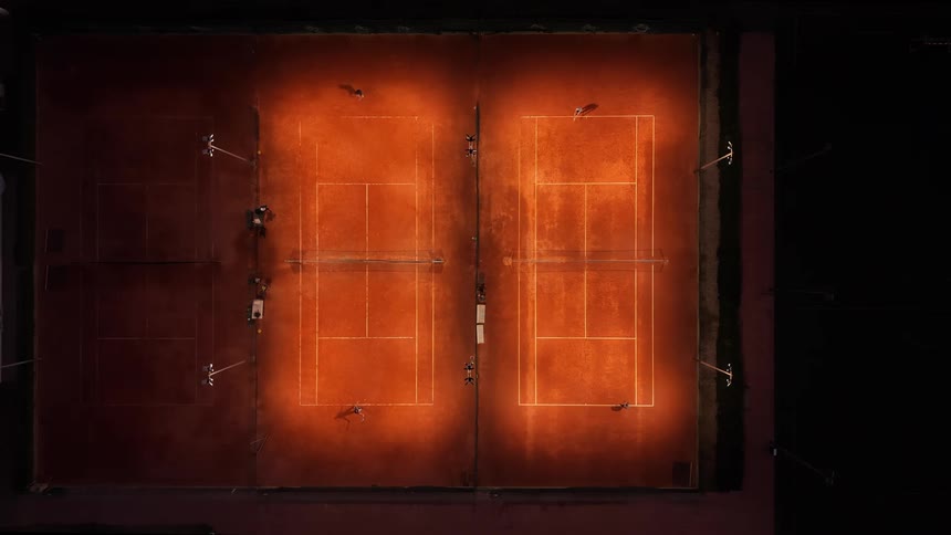 An aerial view captures people playing tennis on illuminated clay courts at night