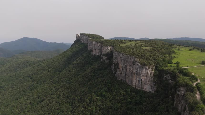 A wide drone shot captures a massive limestone cliff rising above a dense green forest under a hazy sky