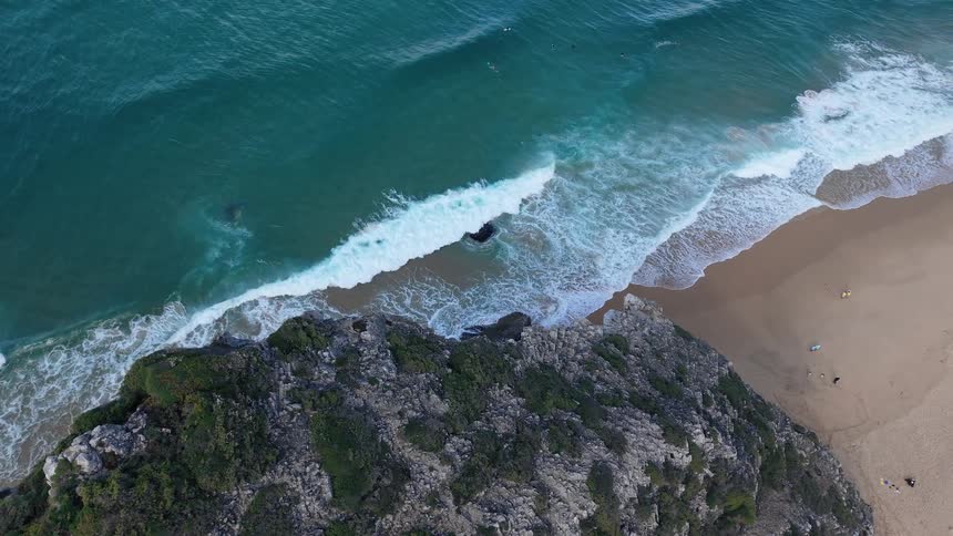 An aerial view shows waves crashing against a rocky cliff and a sandy beach where people are surfing in the ocean