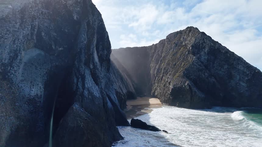 This drone footage captures a dramatic coastal landscape featuring a narrow sandy cove tucked between towering rugged rock cliffs as the camera glides forward and pans sunlight filters through the mist above the beach while white waves crash against th