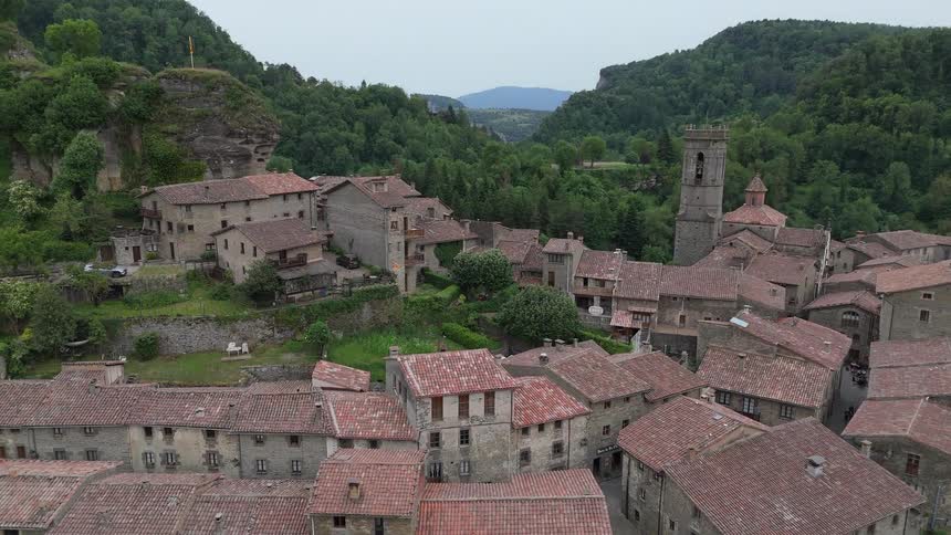 An aerial view reveals a historic stone village with terracotta roofs nestled into the side of a lush forested mountain