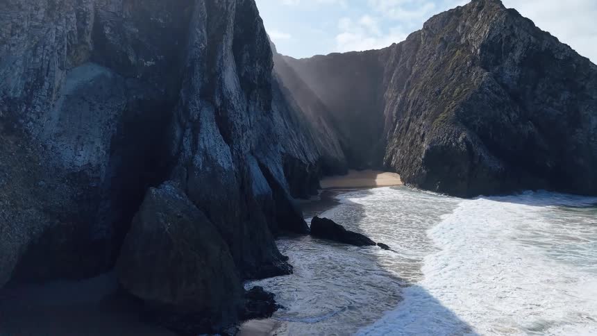 Sunlight streams through a natural arch in a towering cliff as waves crash against the rocky coastline