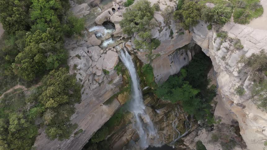 An aerial shot rises to reveal a waterfall cascading down a sheer cliff face into a deep pool below