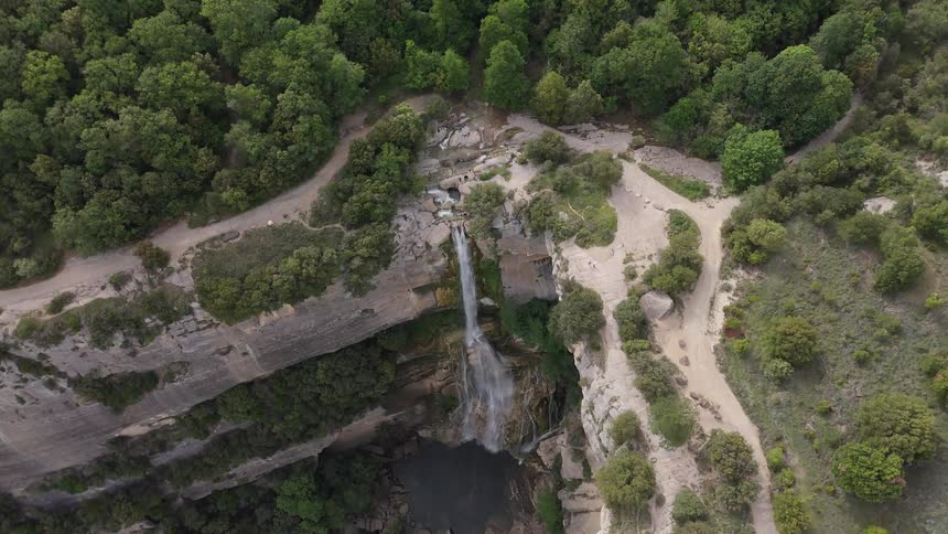 A high altitude drone camera circles above a thin waterfall cascading over a massive curved cliff into a dark pool surrounded by lush green forests and winding dirt trails where a person walks along the rocky precipice