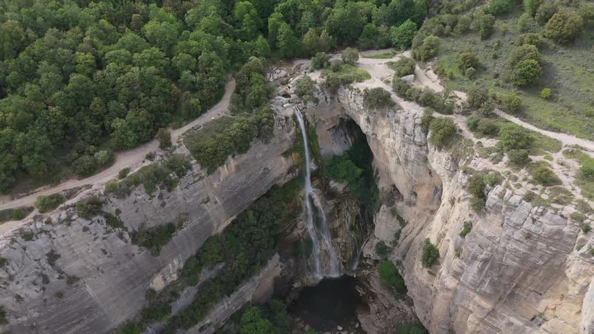 From high above a stunning waterfall cascades from a dense forest plunging down the face of a sheer rock gorge into a dark pool below as tiny hikers walk the cliffside trails