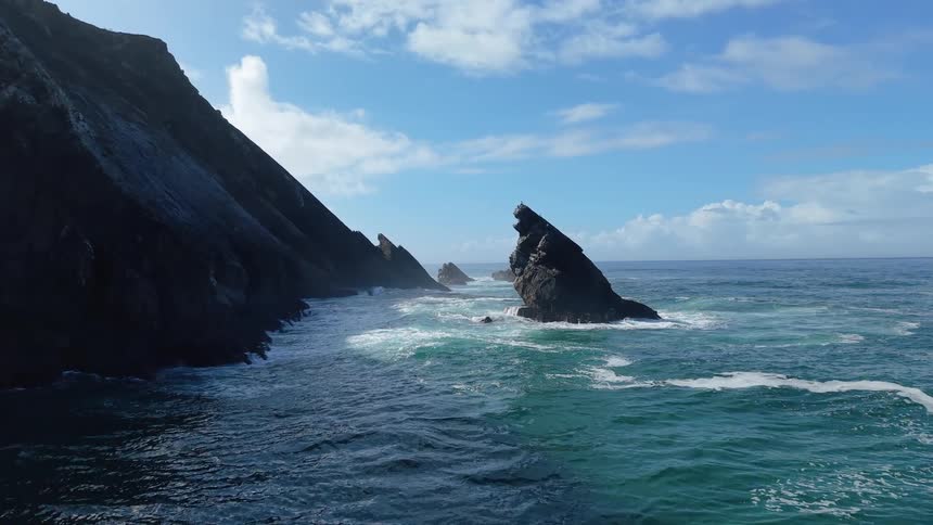 A drone flies over turquoise ocean waves crashing against dark jagged coastal cliffs and sharp rock formations under a blue sky with scattered clouds as a single bird flies through the frame near the steep mountainous shoreline
