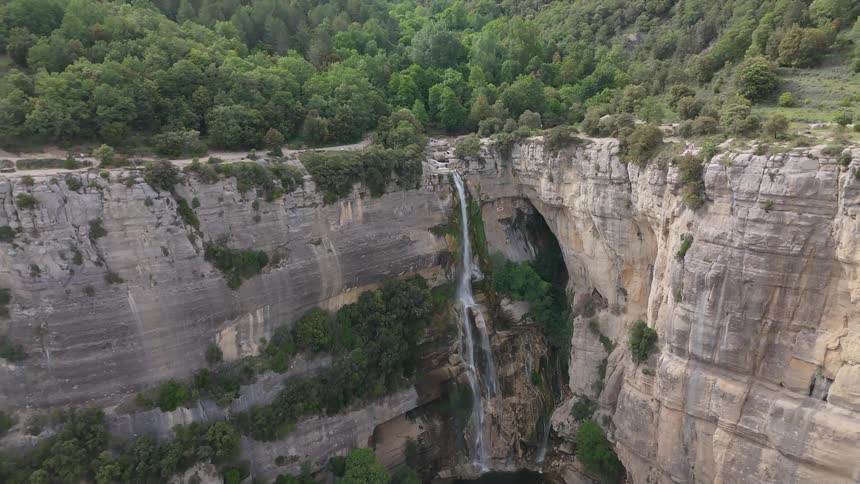 A high angle view shows a thin waterfall cascading down a tall rocky cliff into a pool below surrounded by a lush green forest on a cloudy day